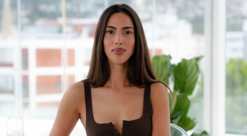 Eine Kandidatin von Die Bachelors 2025 sitzt vor einem Fenster mit Stadtblick und trägt ein braunes, tief ausgeschnittenes Top.