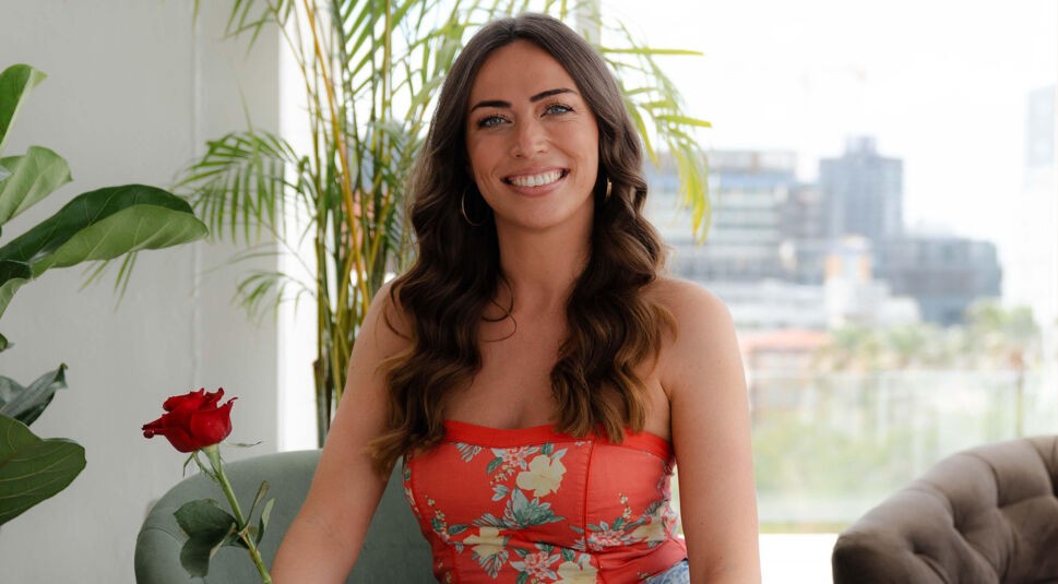 Eine Kandidatin von Die Bachelors 2025 sitzt vor einer Fensterfront mit Stadtblick, trägt ein rotes, geblümtes Top und hält eine rote Rose.