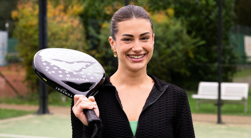 Eine Kandidatin von Die Bachelors 2025 steht auf einem Tennisplatz, trägt eine schwarze Jacke und hält einen schwarzen Padel-Schläger.