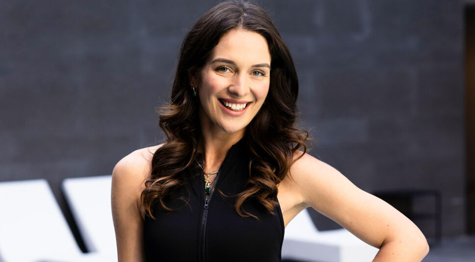 Eine Kandidatin von Die Bachelors 2025 trägt ein schwarzes, ärmelloses Top mit Reißverschluss und hat lockige, braune Haare, im Hintergrund moderne Sitzmöbel.