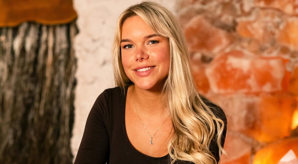 Eine Kandidatin von Die Bachelors 2025 sitzt vor einer rustikalen Steinwand, trägt ein schwarzes Oberteil und hat lange, blonde Haare.