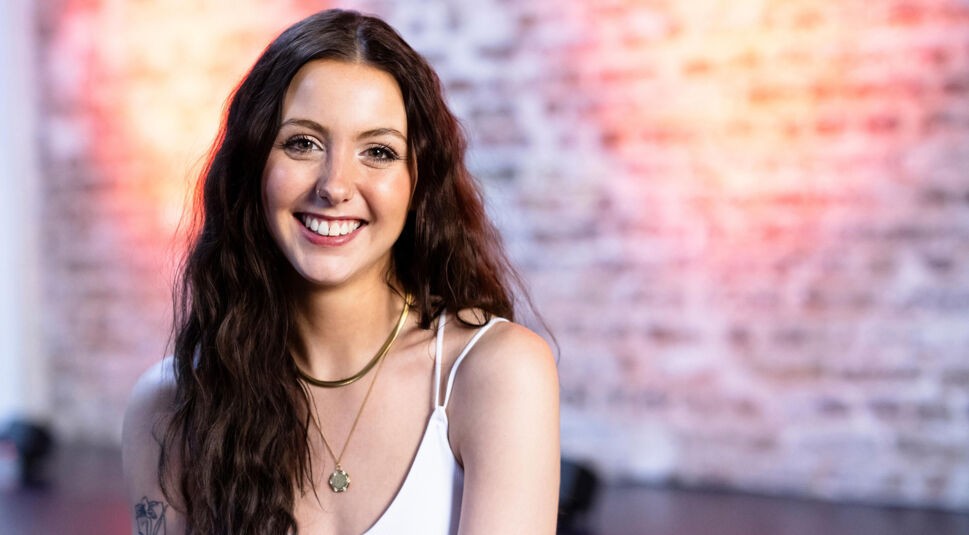 Eine Kandidatin von Die Bachelors 2025 trägt ein weißes Spaghettitop, hat lange, dunkelbraune Haare und steht vor einer Ziegelwand mit Lichtakzenten.