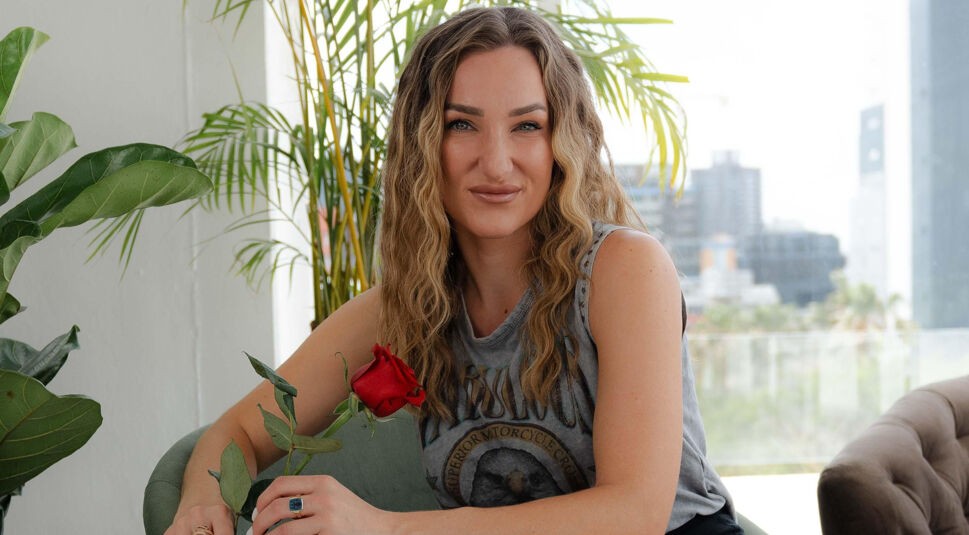 Eine Kandidatin von Die Bachelors 2025 sitzt vor einer Fensterfront mit Skyline, trägt ein graues Printshirt ohne Ärmel und hält eine rote Rose.