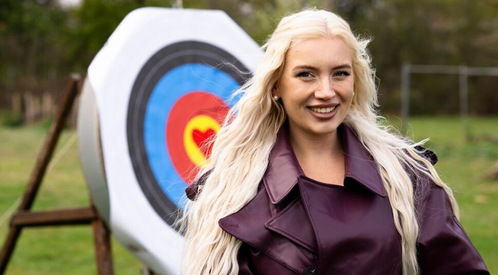 Eine Kandidatin von Die Bachelors 2025 steht im Freien, trägt eine bordeauxfarbene Lederjacke und hat lange, platinblonde Haare.