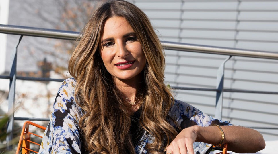 Eine Kandidatin von Die Bachelors 2025 sitzt draußen auf einem orangefarbenen Stuhl, trägt ein gemustertes Oberteil mit Blautönen und hat lange, gewellte Haare.