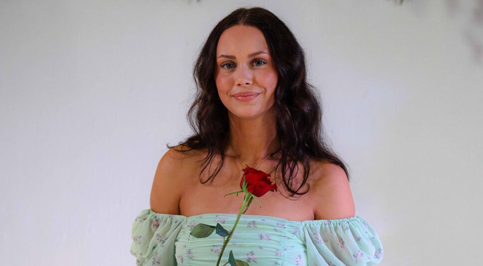 Eine Kandidatin von Die Bachelors 2025 steht vor weißem Hintergrund mit grünen Blättern, trägt ein schulterfreies, mintgrünes Oberteil mit Blumenmuster und hält eine Rose.