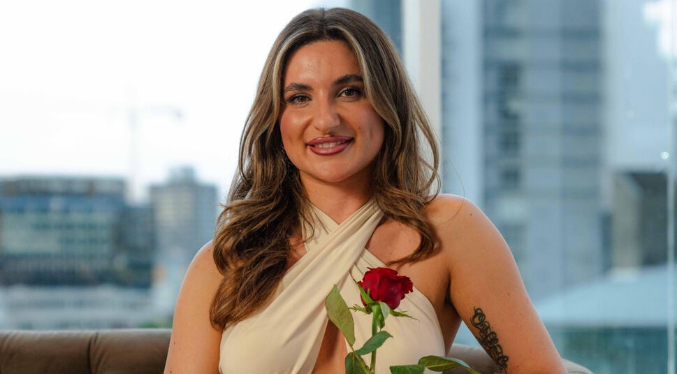 Eine Kandidatin von Die Bachelors 2025 trägt ein cremefarbenes Neckholder-Oberteil, hält eine rote Rose und sitzt vor einer Fensterfront mit Stadtblick.