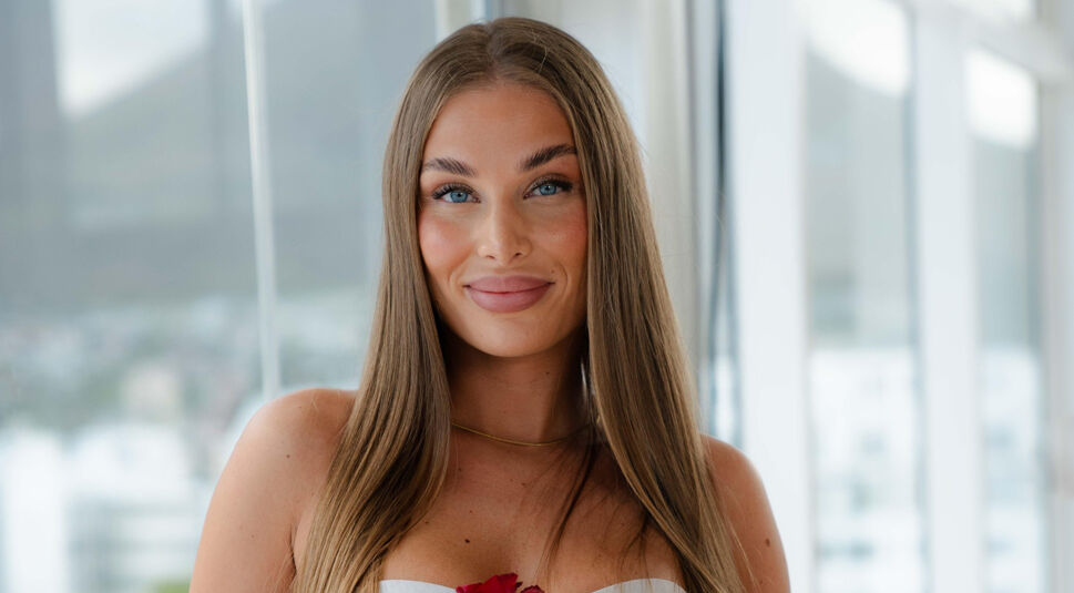 Eine Kandidatin von Die Bachelors 2025 steht vor einer Fensterfront, trägt ein weißes, schulterfreies Kleid und hält eine Rose.