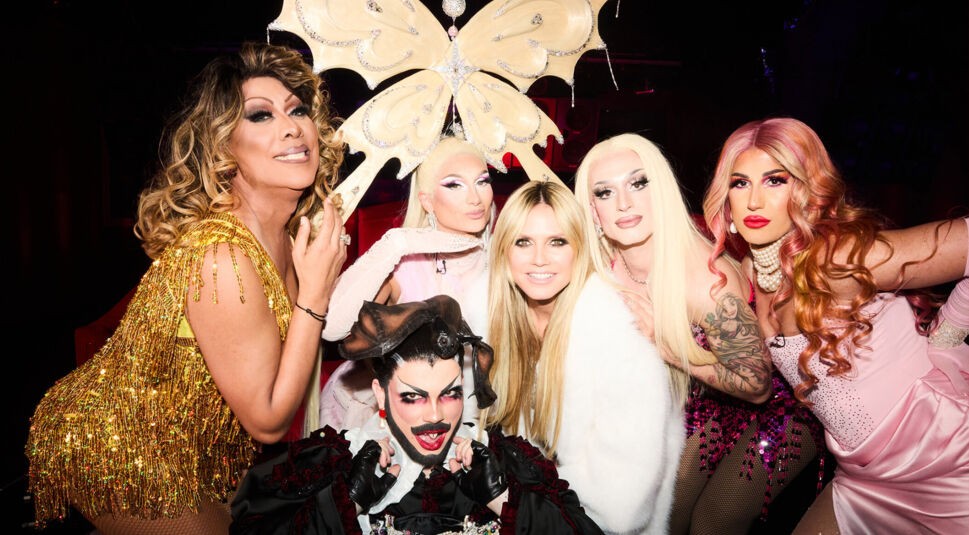 Auf dem Bild zum GNTM 2025 Ticker sind Heidi Klum und fünf bekannte Drag-Performer:innen zu sehen: Yoncé Banks, Katy Bähm, Candy Crash, Bambi Mercury und Catherine Leclery. Sie posieren gemeinsam vor dunklem Hintergrund, teils in glamourösen Gold-, Pink- und Weißtönen. Die Stimmung ist ausgelassen.