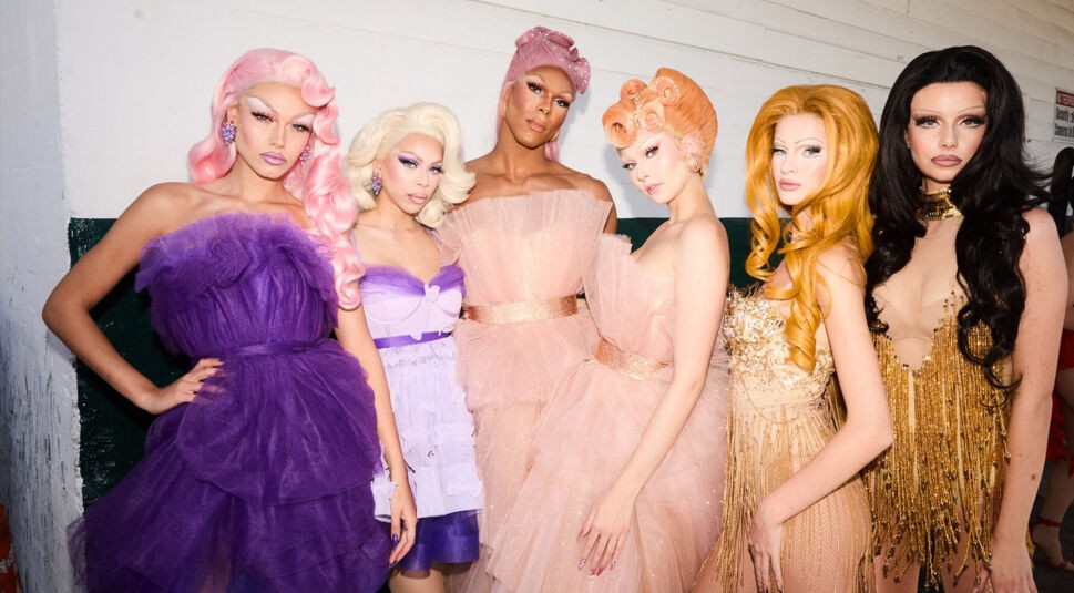 Auf dem Bild zum GNTM 2025 Ticker sind sechs Kandidat:innen zu sehen, gestylt als Drag Queens in auffälligen Tüll- und Glitzerkleidern. Von links nach rechts stehen Daniela (20, Ostfildern), Josy (19, München), Ray (30, Hamburg), Magdalena (21, Wien), Zoe (19, Pulheim) und Katharina (24, Wien). Die Stimmung ist glamourös, alle blicken selbstbewusst in die Kamera, posieren im Backstagebereich einer Modenschau.