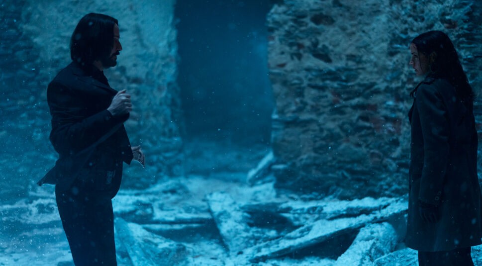 In dieser Szene aus dem Film „Ballerina“, die zur John Wick Ballerina Kritik gehört, steht Keanu Reeves als John Wick links im Bild in dunkler Kleidung und greift mit ernster Miene unter sein Jackett. Rechts steht Ana de Armas als Eve in einem dunklen Mantel. Beide befinden sich in einer verschneiten, ruinösen Umgebung mit angespannten Blicken.