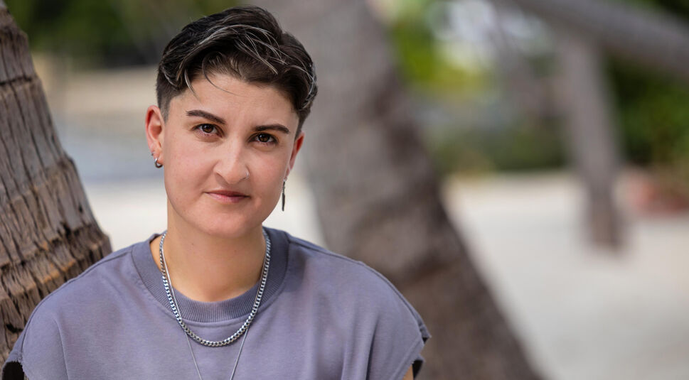 Auf dem Bild in dem Artikel zu Princess Charming 2025 sitzt eine Person mit kurzem, dunklem Haar und grauem T-Shirt an einer Palme.