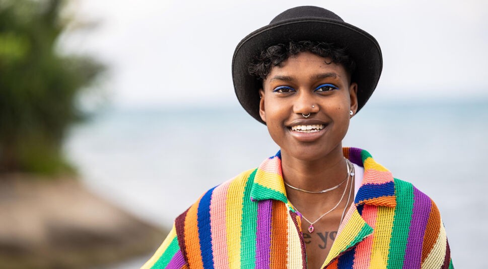 Auf dem Bild in dem Artikel zu Princess Charming 2025 steht eine Person mit bunter Jacke und schwarzem Hut am Meer.