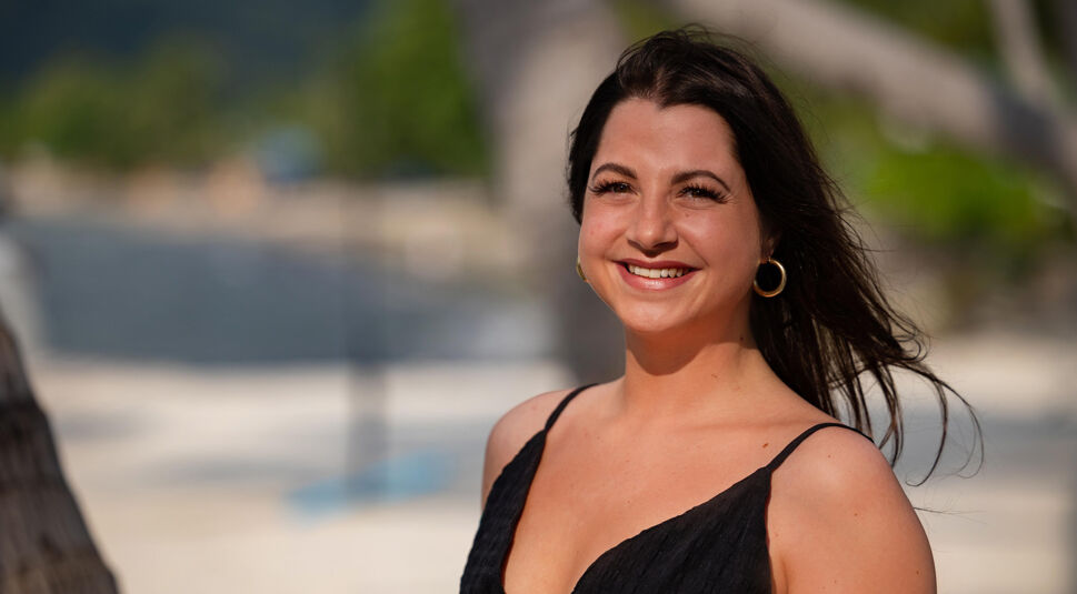 Auf dem Bild in dem Artikel zu Princess Charming 2025 steht eine Person im schwarzen Kleid mit dunklen Haaren am Strand.