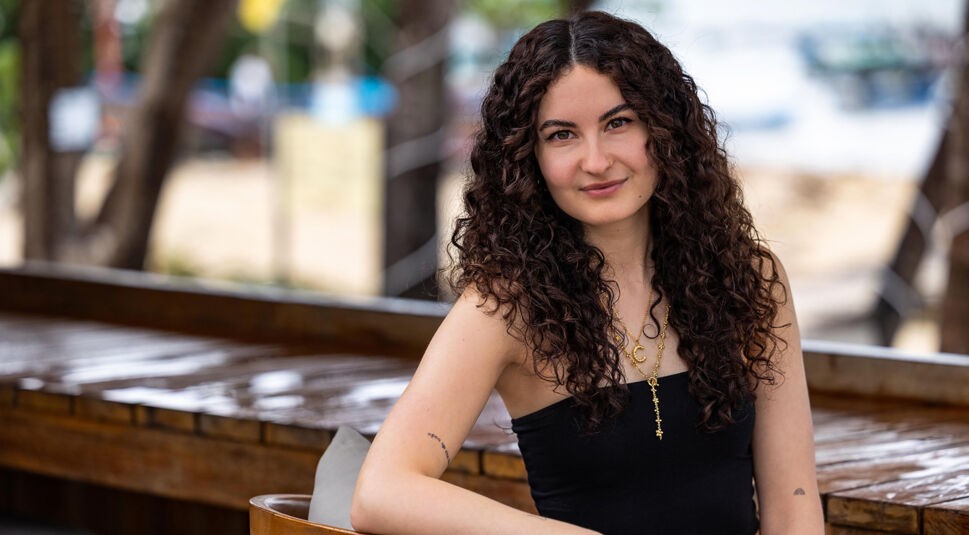 Auf dem Bild in dem Artikel zu Princess Charming 2025 sitzt eine Person mit lockigen Haaren an einem Holztresen.