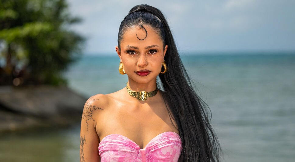 Auf dem Bild in dem Artikel zu Princess Charming 2025 steht eine Person in pinkem Kleid mit Goldschmuck am Strand.