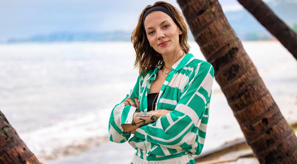 Auf dem Bild in dem Artikel zu Princess Charming 2025 steht eine Person in grün-weißem Outfit zwischen Palmen am Strand.