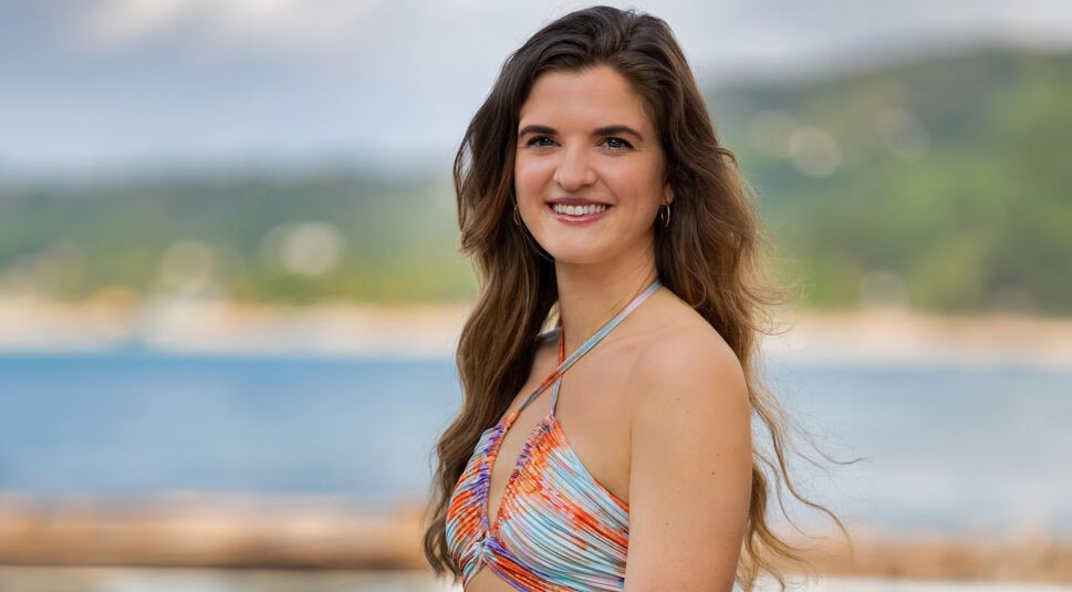 Auf dem Bild in dem Artikel zu Princess Charming 2025 steht eine Person mit langen, braunen Haaren in einem bunten Neckholder-Top am Strand.