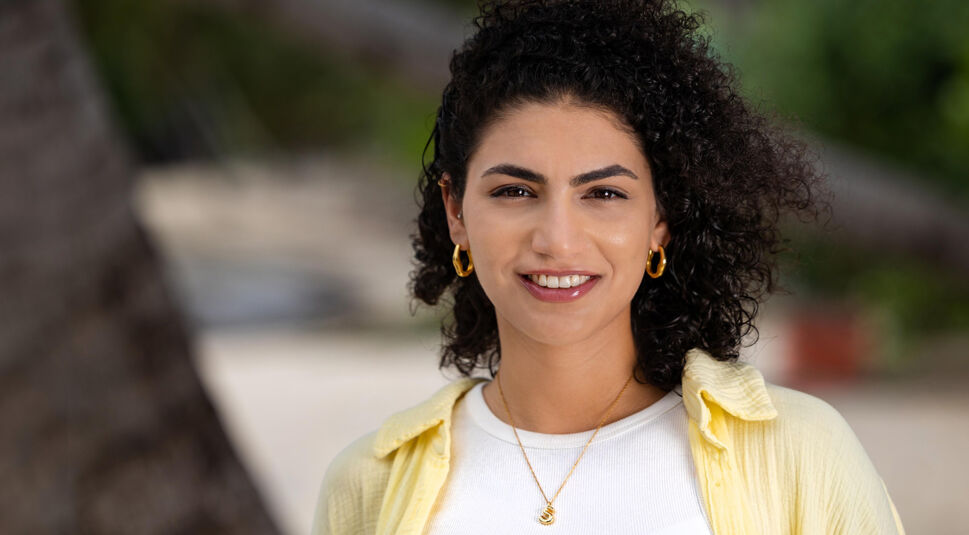 Auf dem Bild in dem Artikel zu Princess Charming 2025 steht eine Person mit lockigem, dunklem Haar, weißem Shirt und gelbem Hemd vor Palmen am Strand.