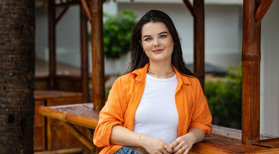 Auf dem Bild in dem Artikel zu Princess Charming 2025 lehnt eine Person mit langen, dunklen Haaren, weißem Top und orangener Bluse an einer Holzstange.