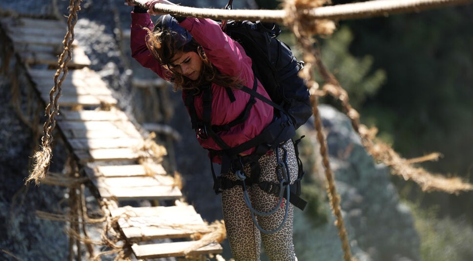 In dieser Szene aus der Reality-Show The Summit überquert eine Teilnehmerin konzentriert eine wackelige Hängebrücke aus Holz und Seilen. Sie trägt einen Klettergurt, einen großen Rucksack und hält sich mit beiden Händen an den Seilen fest. Die Herausforderung wirkt körperlich fordernd und der Moment ist angespannt und abenteuerlich.