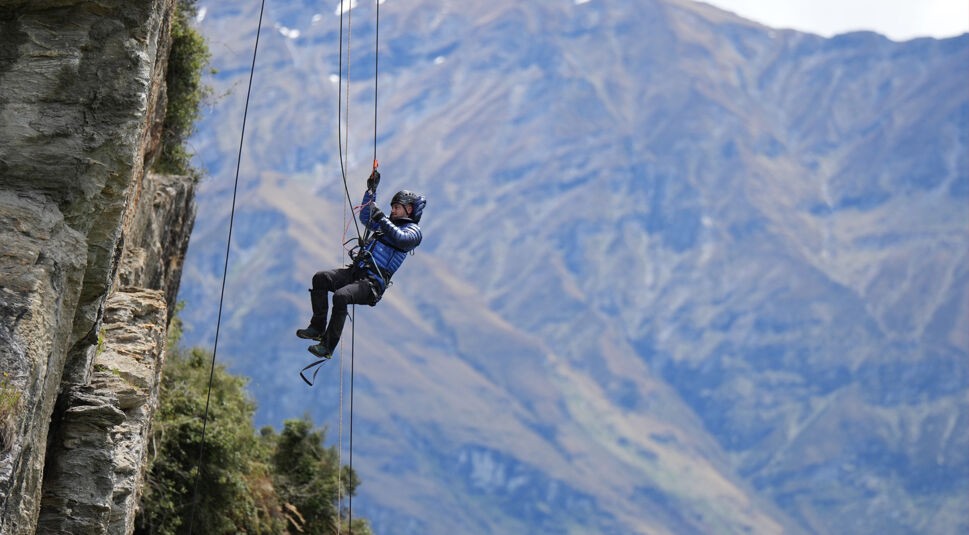 In einer Szene aus der Reality-Show The Summit hängt ein Teilnehmer gesichert mit Kletterseilen an einer steilen Felswand. Er trägt Schutzhelm und Outdoor-Kleidung, während im Hintergrund majestätische Bergketten zu sehen sind. Die Stimmung ist angespannt und abenteuerlich, typisch für die Herausforderungen in The Summit.