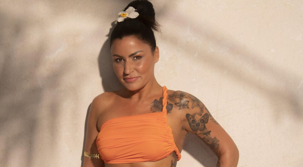 Auf dem Bild zur Villa der Versuchung steht Jasmin Herren vor einer beigen Wand. Sie trägt ein schulterfreies orangefarbenes Top mit geflochtenem Träger und eine weiße Blume im hochgesteckten Haar.