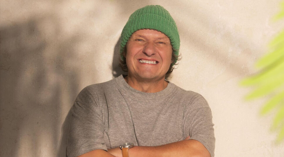 Auf dem Bild zur Villa der Versuchung ist Manni Ludolf vor einer hellen Wand zu sehen. Er trägt ein graues T-Shirt, eine grüne Strickmütze und eine Armbanduhr mit braunem Band. Seine Arme sind verschränkt und er lächelt breit in die Kamera.