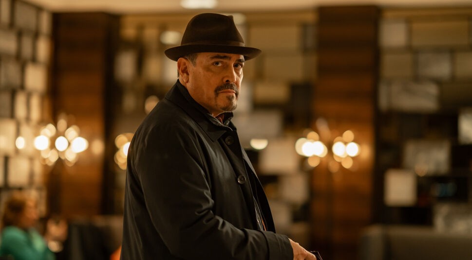 Auf dem Bild zur möglichen Dexter: Wiedererwachen Staffel 2 ist Angel Batista zu sehen, gespielt von David Zayas. Er trägt einen dunklen Mantel und einen braunen Hut, während er sich mit ernstem Blick über die Schulter dreht. Die warme Beleuchtung und das elegante Ambiente im Hintergrund erzeugen eine geheimnisvolle und spannungsgeladene Atmosphäre.