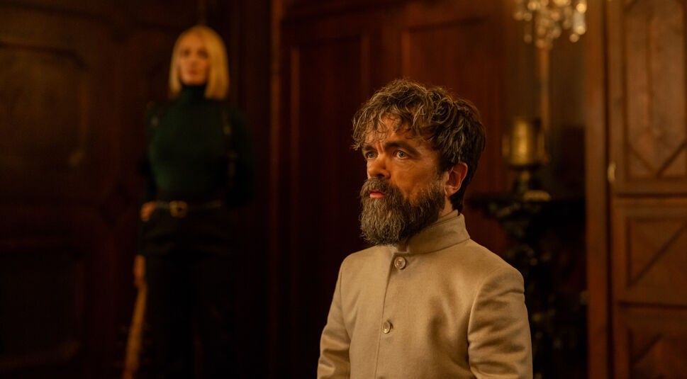 Auf dem Bild zur Dexter: Wiedererwachen Staffel 2 ist Leon Prater zu sehen, gespielt von Peter Dinklage. Er kniet in einem eleganten Raum mit dunkler Holzverkleidung und blickt konzentriert nach vorn. Im Hintergrund steht Charley, dargestellt von Uma Thurman, unscharf aber wachsam. Die Szene wirkt angespannt und voller Dramatik.