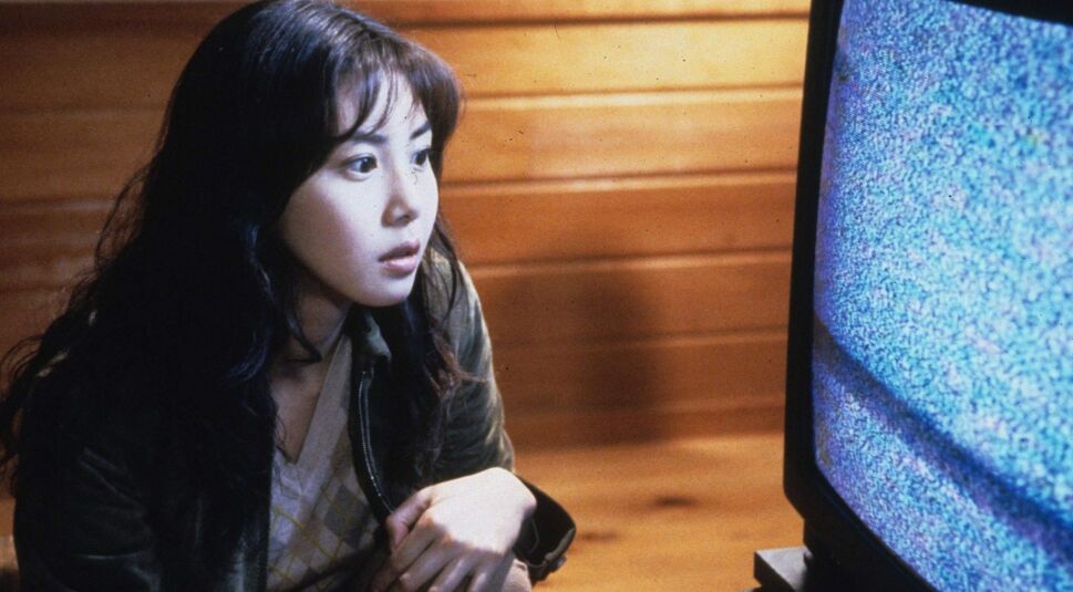 In den GigaTV-Highlights im Oktober 2025 ist eine Szene aus dem japanischen Horrorfilm „Ringu“ zu sehen. Eine junge Frau sitzt in einem holzvertäfelten Raum vor einem Röhrenfernseher, der ein stark verrauschtes Bild zeigt.
