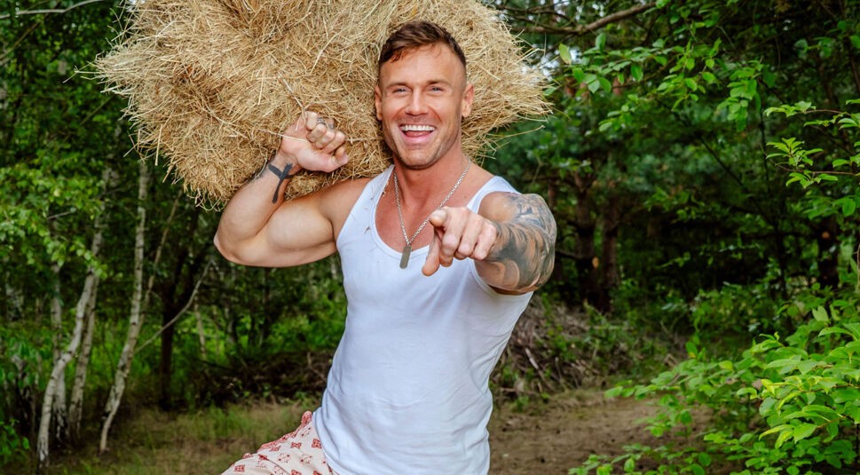 Auf dem Bild zur Sendung Der Promihof trägt Chris ein Heubündel lässig auf der Schulter und zeigt mit einem breiten Grinsen in die Kamera. Er trägt ein weißes Tanktop, eine gemusterte Hose und zeigt stolz seine tätowierten Arme. Die Szene wirkt dynamisch, sympathisch und betont seine sportliche Ausstrahlung in der ländlichen Umgebung.