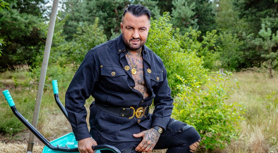 Auf dem Bild zur Show Der Promihof sitzt Cosimo in auffälligem Look auf einer umgedrehten Schubkarre vor grüner Waldkulisse. Er trägt ein schwarzes Outfit mit goldenen Knöpfen und auffälligem Gürtel, zeigt tätowierte Brust und Schmuck. Sein Blick ist direkt und selbstbewusst – die Szene wirkt extravagant und ländlich zugleich.