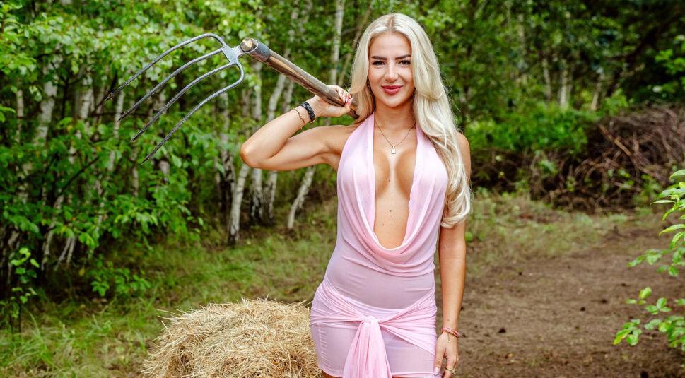 Auf dem Bild zur Show Der Promihof steht Emma mit einer Heugabel lässig über der Schulter vor einer grünen Waldkulisse. Sie trägt ein figurbetontes, rosafarbenes Kleid mit tiefem Ausschnitt und posiert selbstbewusst neben einem Heuballen. Die Szene verbindet ländliche Umgebung mit auffälligem Glamour und starker Präsenz.