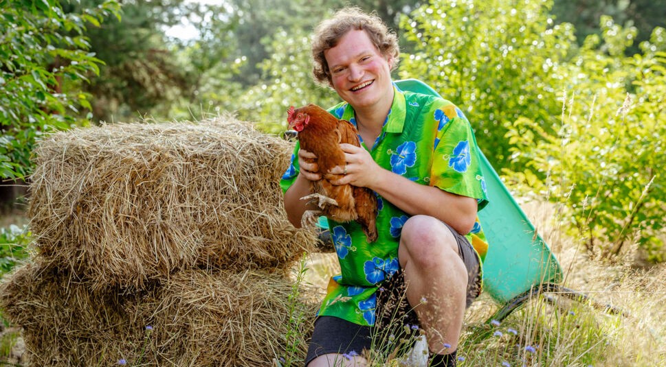 Auf dem Bild zur Show Der Promihof kniet Fritz lachend im hohen Gras und hält ein braunes Huhn liebevoll im Arm. Er trägt ein buntes Hawaiihemd mit blauen Blumen und sitzt vor zwei Heuballen und einer grünen Schubkarre. Die Szene wirkt herzlich, bodenständig und voller ländlichem Charme.