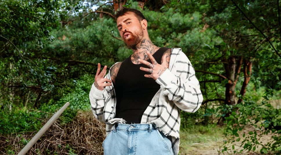 Auf dem Bild zur Sendung Der Promihof steht Gigi mit cooler Pose im Wald, beide Hände zeigen lässige Gesten. Er trägt ein schwarzes Tanktop, eine offene kariert-beige Jacke und helle Jeans. Seine Tattoos am Hals und an den Händen sind deutlich sichtbar. Die Szene wirkt rebellisch, authentisch und urban im Kontrast zur Naturkulisse.