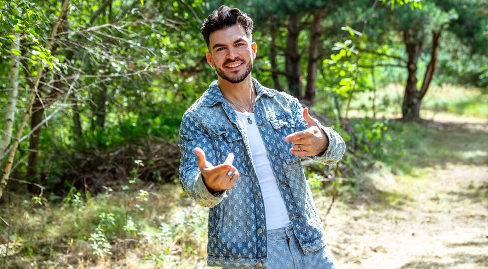Auf dem Bild zur Show Der Promihof steht Maki lässig inmitten einer sonnigen Waldlichtung und zeigt mit beiden Händen in die Kamera. Er trägt ein auffälliges Jeans-Outfit mit Muster, bestehend aus Jacke und Hose, kombiniert mit einem weißen Shirt. Sein charismatisches Lächeln und seine lockere Ausstrahlung prägen die lebendige Szene.