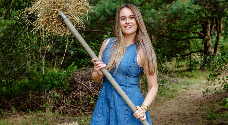 Auf dem Bild zur Show Der Promihof hält Nina eine Heugabel mit frischem Heu in der Hand und lächelt in die Kamera. Sie trägt ein ärmelloses Jeanskleid und steht vor einer waldigen Kulisse. Der Look ist natürlich und bodenständig, die Szene strahlt sommerliche Leichtigkeit und ländlichen Charme aus.