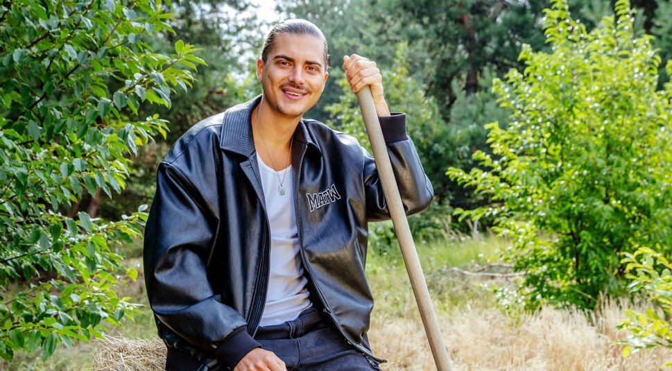 Auf dem Bild zur Sendung Der Promihof sitzt Tim lässig auf einem Heuballen und lehnt sich an eine Heugabel. Er trägt eine schwarze Lederjacke mit dem Schriftzug „MAHN“, ein weißes Tanktop und dunkle Jeans. Sein zurückgegeltes Haar und das selbstsichere Lächeln verleihen der Szene eine coole, urbane Note inmitten ländlicher Kulisse.