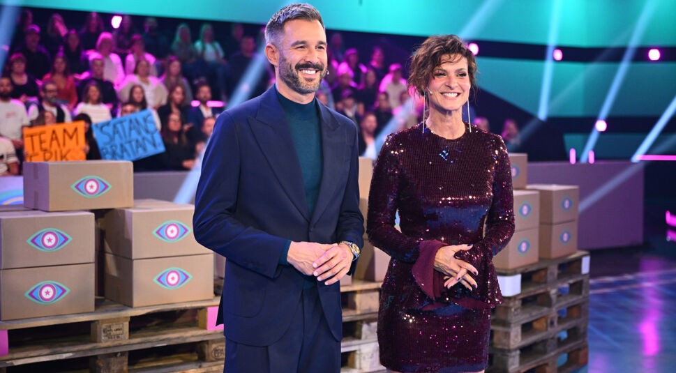 In dieser Szene aus „Promi Big Brother“ 2025 stehen die beiden Moderator:innen lächelnd im Studio.