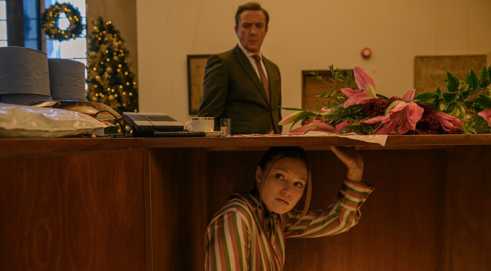 In dieser Szene aus dem Netflix-Weihnachtsfilm Jingle Bell Heist versteckt sich Olivia Holt als Sophia unter einem Schreibtisch, während ihr Chef Maxwell Sterling, gespielt von Peter Serafinowicz, ahnungslos darüber steht. Die Szene wirkt angespannt und humorvoll zugleich – auf dem Tisch liegen auffällige Blumen, während im Hintergrund ein festlich geschmückter Weihnachtsbaum leuchtet.