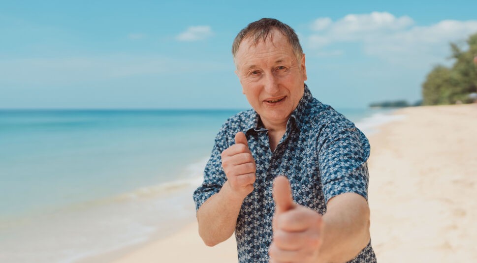 Auf dem Bild zu Kampf der RealityAllstars 2026 ist Schäfer Heinrich am Strand zu sehen. Er steht nah an der Kamera, lächelt freundlich und zeigt einen Daumen nach oben, während er mit der anderen Hand eine lockere Faust macht. Im Hintergrund erstrecken sich Meer und heller Sandstrand unter blauem Himmel, was eine entspannte, sommerliche und positive Stimmung vermittelt.