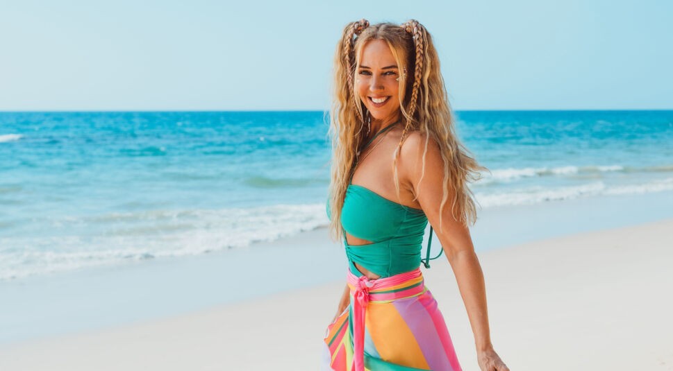 Auf dem Bild zu Kampf der RealityAllstars 2026 ist Loona am Strand zu sehen. Sie steht leicht seitlich zur Kamera, lächelt strahlend und trägt ein auffälliges Outfit mit grünem Oberteil und buntem, mehrfarbigem Rock. Ihre geflochtenen Zöpfe unterstreichen den sommerlichen Look, während das Meer im Hintergrund eine warme, lebendige Urlaubsstimmung erzeugt.
