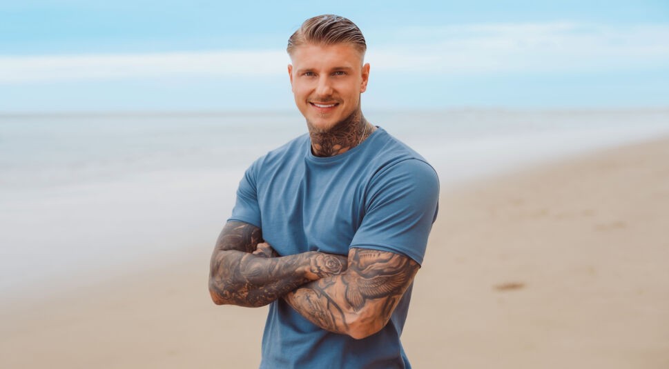 Auf dem Bild zu Kampf der RealityAllstars 2026 ist Paco Herb am Strand zu sehen. Er steht mit verschränkten Armen vor der Kamera, lächelt selbstbewusst und trägt ein schlichtes, blaues T-Shirt. Seine auffälligen Tattoos an Armen und Hals unterstreichen seinen markanten Look, während das Meer im Hintergrund eine ruhige, sommerliche Atmosphäre schafft.