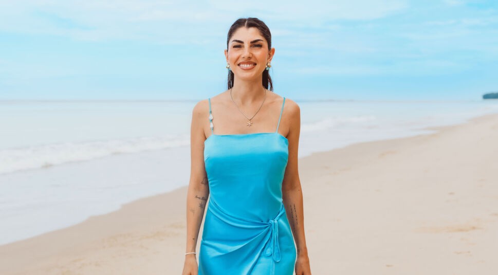 Auf dem Bild zu Kampf der RealityAllstars 2026 ist Yeliz Koc am Strand zu sehen. Sie steht frontal zur Kamera, lächelt selbstbewusst und trägt ein eng anliegendes, türkisfarbenes Kleid mit schmalen Trägern. Der ruhige Ozean und der helle Sandstrand im Hintergrund erzeugen eine sommerliche, entspannte und zugleich stilvolle Atmosphäre.