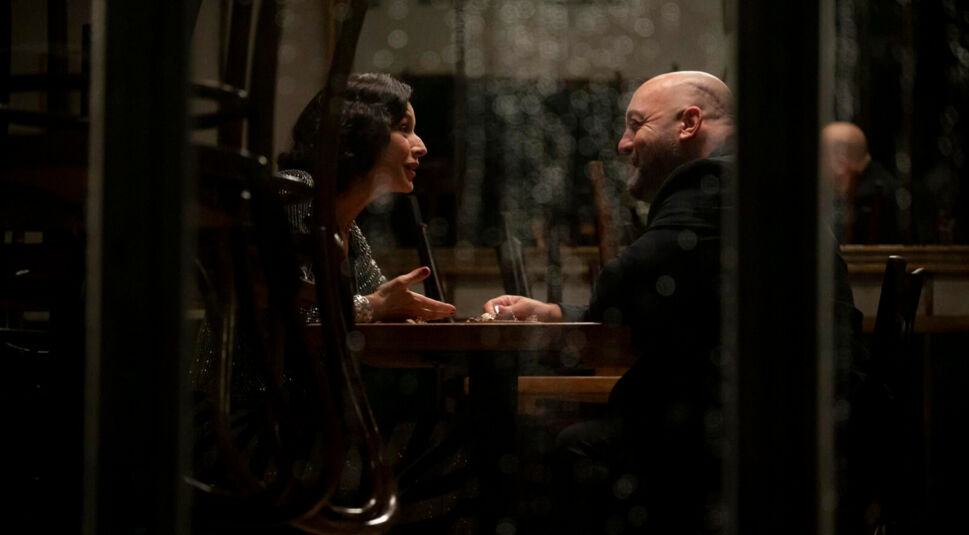 Auf dem Bild aus dem Solo Mio-Film sitzen Gia, gespielt von Nicole Grimaudo, und Matt, gespielt von Kevin James, abends an einem kleinen Tisch in einem geschlossenen Café. Durch eine regennasse Fensterscheibe sieht man, wie sich beide tief in die Augen blicken und lächeln. Die intime Stimmung ist ruhig, romantisch und vertraut.