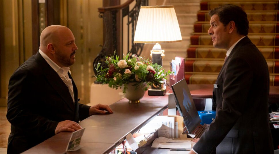 Auf dem Bild zum Film Solo Mio mit Kevin James steht Matt, gespielt von Kevin James, an einer Hotelrezeption und spricht mit einem Hotelrezeptionisten im dunklen Anzug. Die Szene spielt in einer edlen Hotellobby mit Teppichtreppe, Blumengesteck und Tischlampe, was eine gehobene, leicht angespannte Atmosphäre vermittelt.