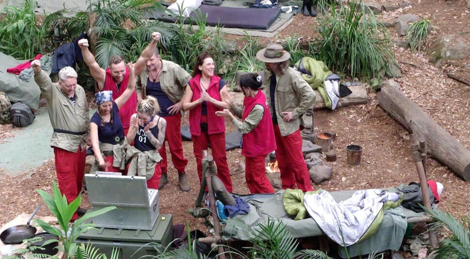 Auf dem Bild zu Dschungelcamp Tag 13 sind mehrere Campbewohner:innen im australischen Dschungel zu sehen, die gemeinsam um eine geöffnete Kiste stehen. Einige jubeln mit erhobenen Armen, andere klatschen oder schauen gespannt hinein. Alle tragen typische rote Dschungelcamp-Hosen. Die Stimmung ist ausgelassen, erleichtert und von gemeinsamer Freude geprägt.