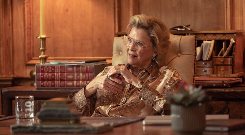 Auf dem Bild zur Dutton Ranch ist Annette Bening als Beulah Jackson in einem elegant eingerichteten Raum zu sehen. Sie sitzt entspannt in einem Sessel, trägt eine gemusterte Bluse und auffälligen Schmuck. Ihr aufmerksamer Blick und die warme Beleuchtung vermitteln eine ruhige, kontrollierte und zugleich leicht dominante Atmosphäre.