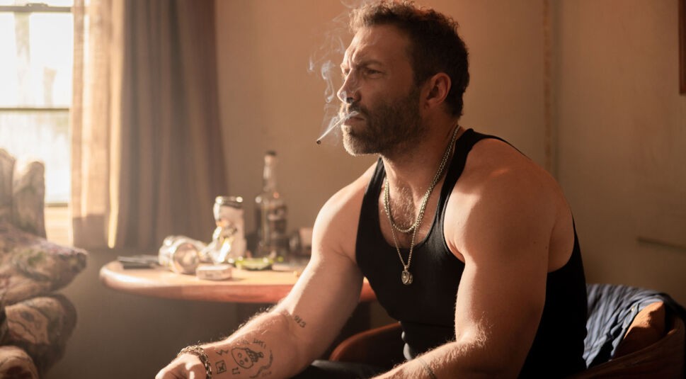 Auf dem Bild zur Dutton Ranch ist Jai Courtney als Rob-Will in einer ruhig-intensiven Szene zu sehen. Er sitzt in einem schwach beleuchteten Raum, trägt ein schwarzes Tanktop und mehrere Ketten, während er eine Zigarette raucht. Sein nachdenklicher Blick und die warme Lichtstimmung verleihen der Szene eine raue, angespannte Atmosphäre.